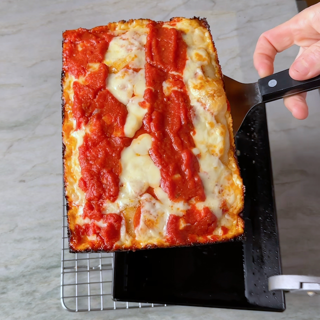 Detroit-Style pizza being transferred from an Ooni Detroit-Style Pizza Pan to a rack to cool.